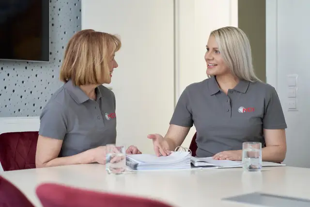 Zwei Kolleginnen im Poloshirt besprechen einen Ordner mit Unterlagen im Besprechungsraum