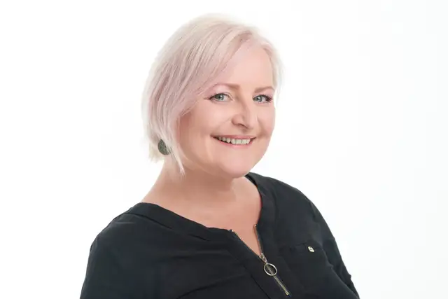 Frau mit rosablondem Bob im schwarzen Oberteil lacht herzlich vor weißem Studiohintergrund, freundliches Business-Portrait