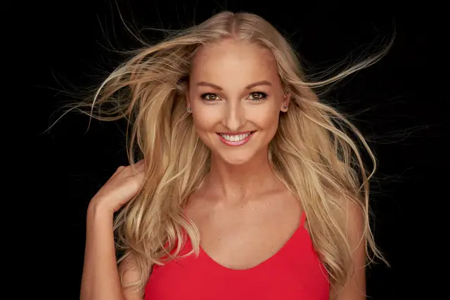 Junge Frau mit wehendem blondem Haar im roten Top strahlt vor schwarzem Hintergrund, ausdrucksstarkes Studio-Portrait