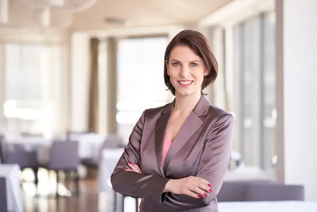 Geschäftsfrau im taupefarbenen Seidenblazer mit verschränkten Armen lächelt in hellem Restaurant, Business-Portrait