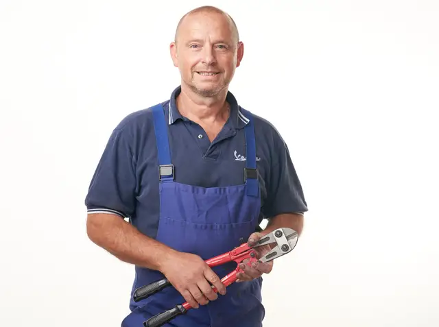 Handwerker in blauer Arbeitslatzhose mit Bolzenschneider lächelt vor weißem Hintergrund, sympathisches Mitarbeiterportrait