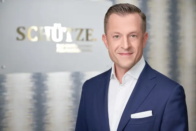 Unternehmer im dunkelblauen Sakko mit weißem Hemd lächelt vor Firmenwand mit goldenem Schriftzug, elegantes Business-Portrait