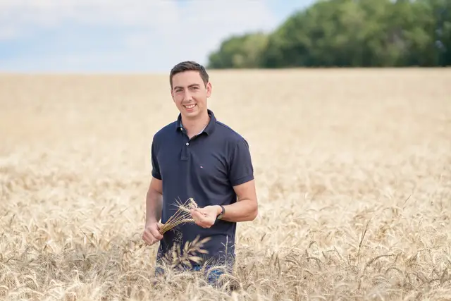 Junger Landwirt im dunkelblauen Polo steht mit Ähren in der Hand im sommerlichen Weizenfeld, authentisches Portrait