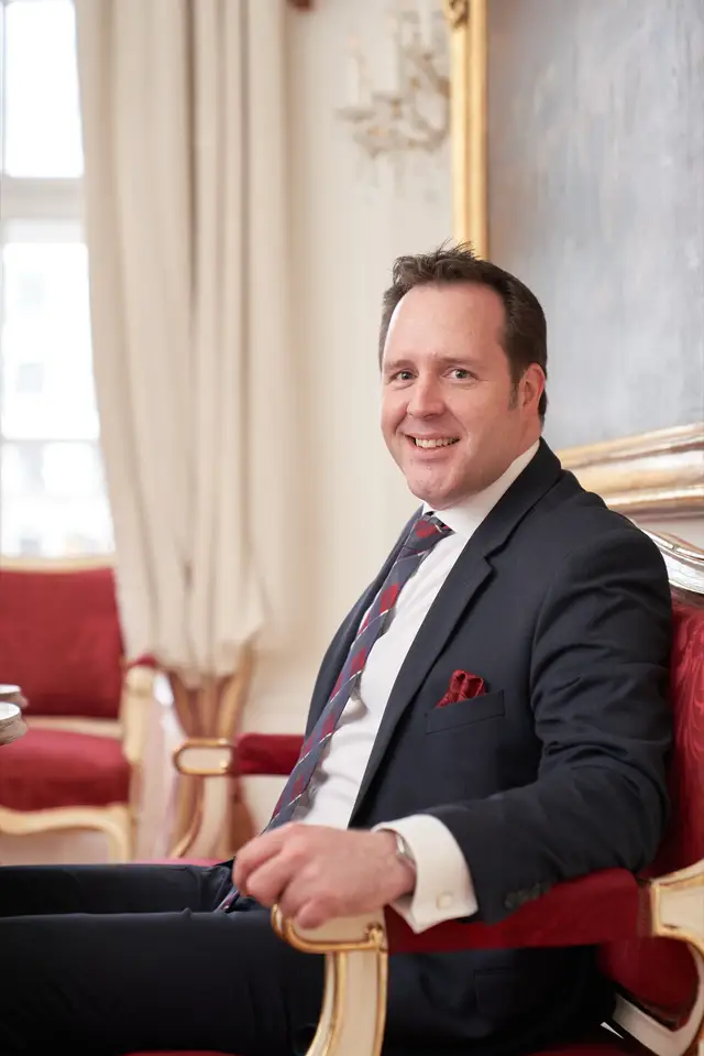 Lächelnder Mann im dunklen Anzug mit Krawatte sitzt auf rotem Barockfauteuil in prunkvollem Salon, klassisches Portrait