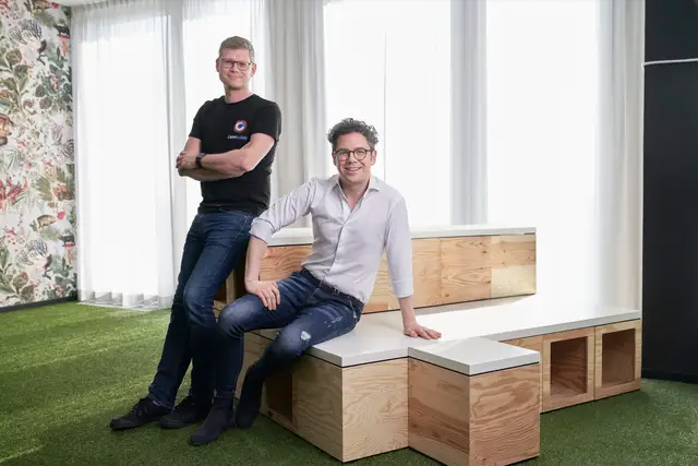 Zwei junge Gründer in legerer Kleidung posieren entspannt auf Holzmöbeln im modernen Büro mit Kunstrasen, Team-Portrait