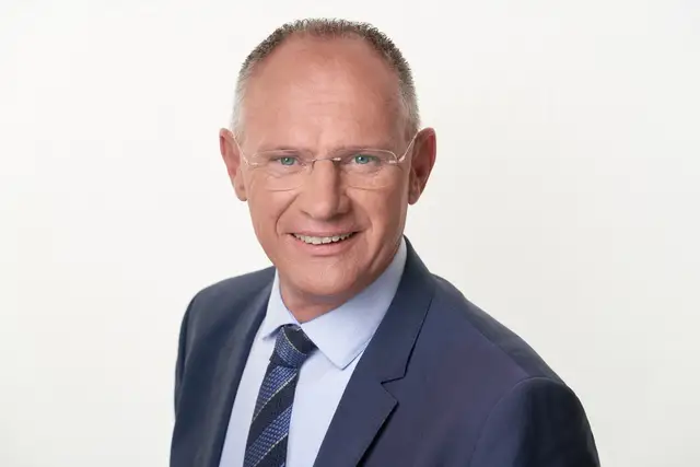 Mann mit randloser Brille im blauen Anzug mit Krawatte lächelt vor hellem Hintergrund, klassisches Business-Portrait