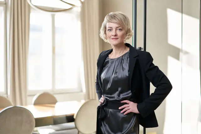 Blonde Frau im schwarzen Blazer über grauem Seidenkleid steht mit Hand in der Hüfte in elegantem Interieur, Portrait