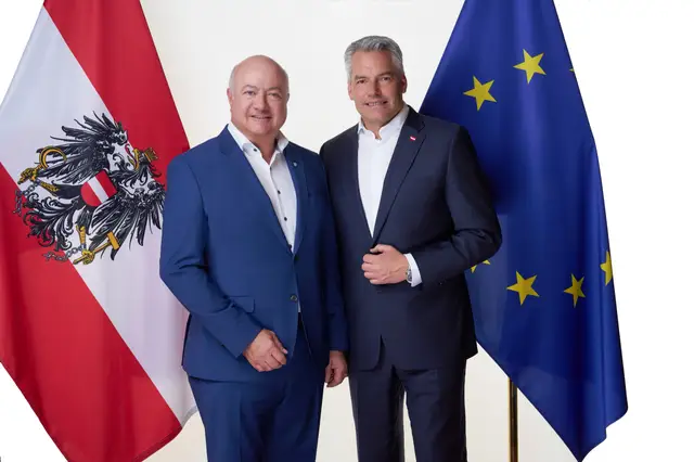 Zwei Männer in dunkelblauen Anzügen stehen lächelnd zwischen Österreich- und EU-Flagge, offizielles Portrait