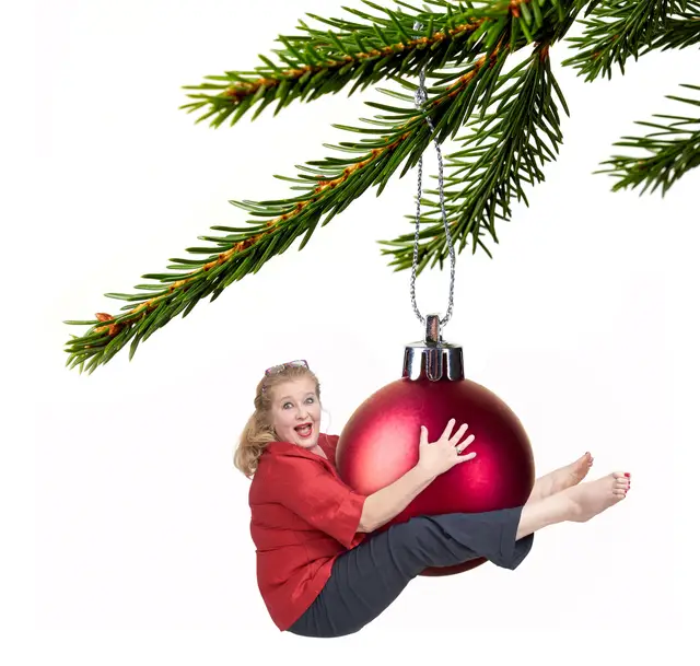 Frau hängt als lebendiger Christbaumschmuck an einer riesigen roten Kugel am Tannenzweig, humorvolle Bildmontage