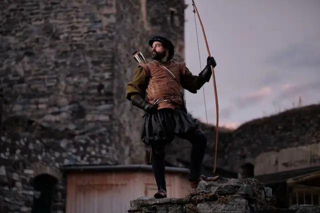 Bogenschütze in historischer Gewandung mit Langbogen auf einer Burgruine, stimmungsvoll inszeniert in der Abenddämmerung